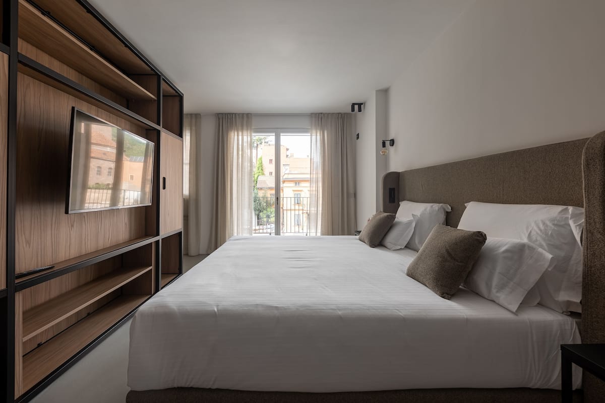 A hotel room with a large bed, a TV mounted on a wooden panel, a balcony with curtains, and a view of buildings.
