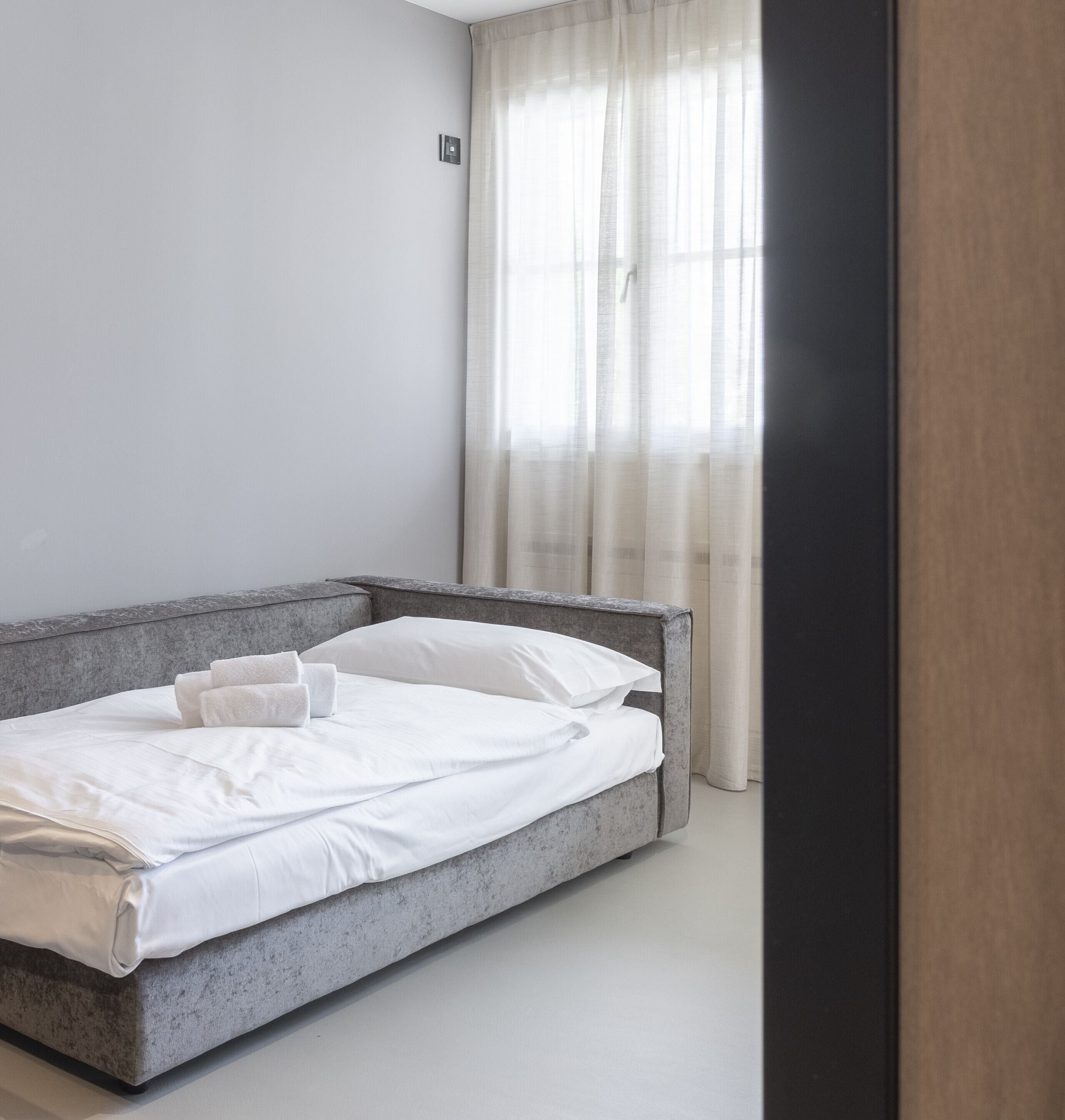 A modern bedroom with a grey bed, white linens, and sheer curtains.