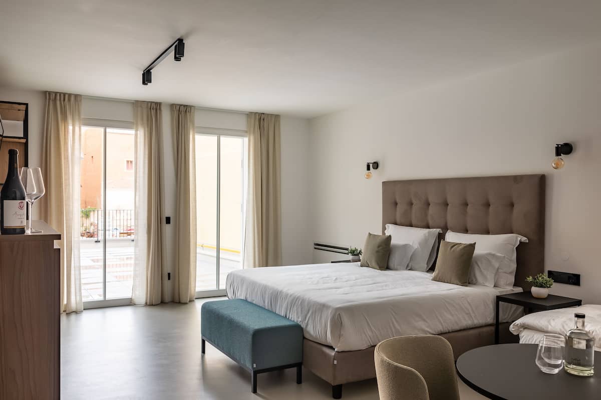 A modern hotel room with a large bed, a bench, a round table, and a view through the glass doors.