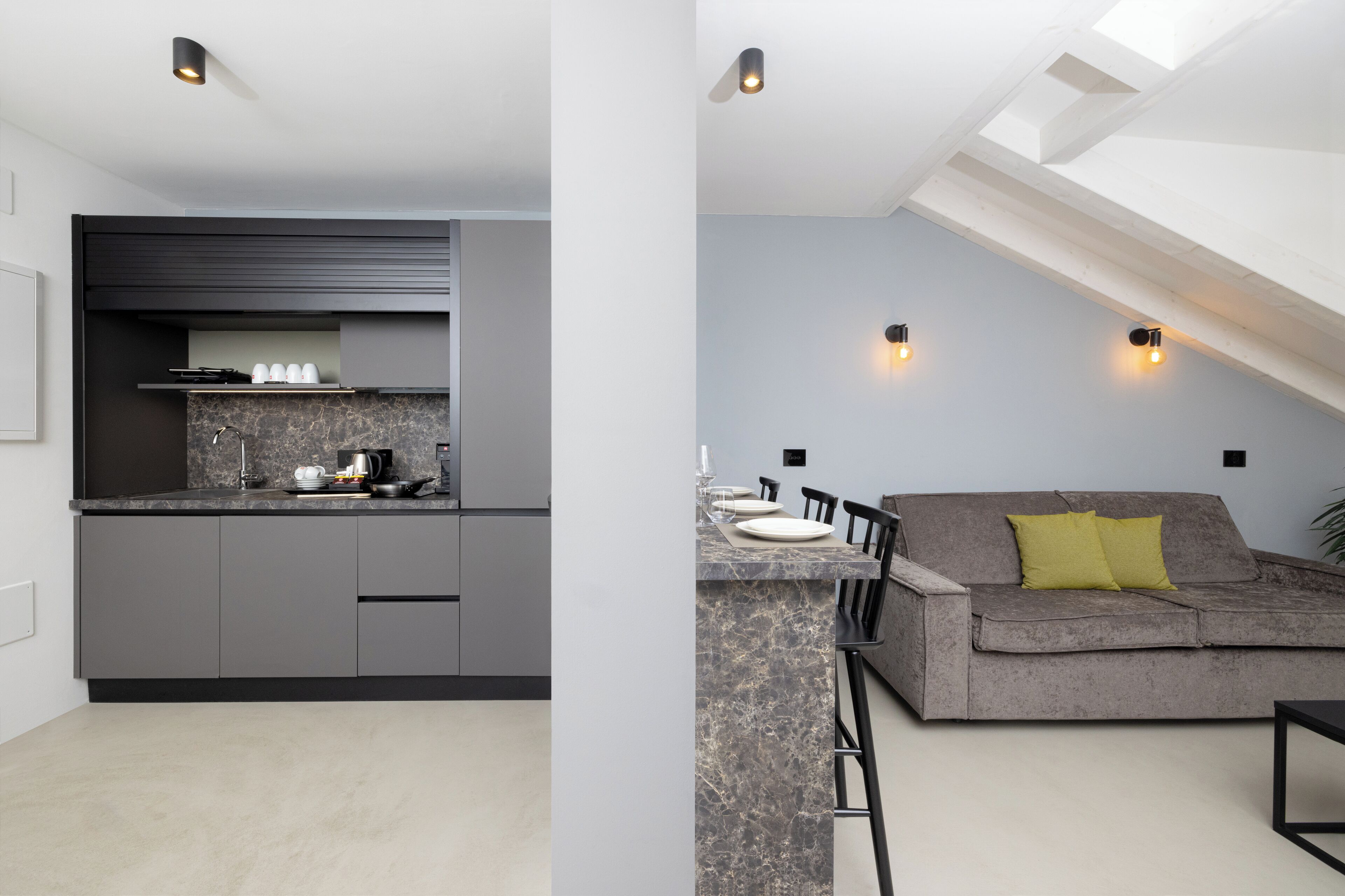 A modern kitchen with grey cabinets and a marble countertop, a dining area with a grey sofa and yellow pillow, and a ceiling with skylights.