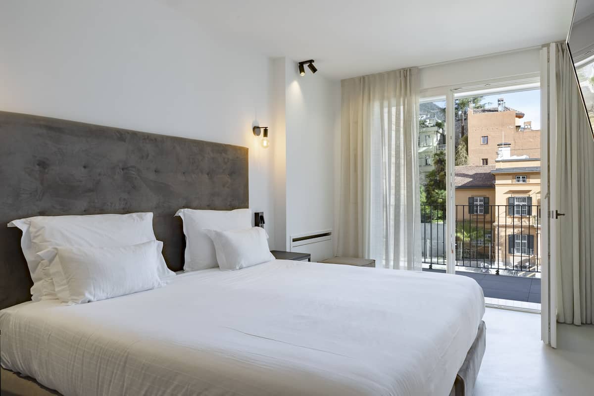 A hotel room with a large bed, a modern headboard, a bedside table, a nightstand, and a view of the city through a balcony.