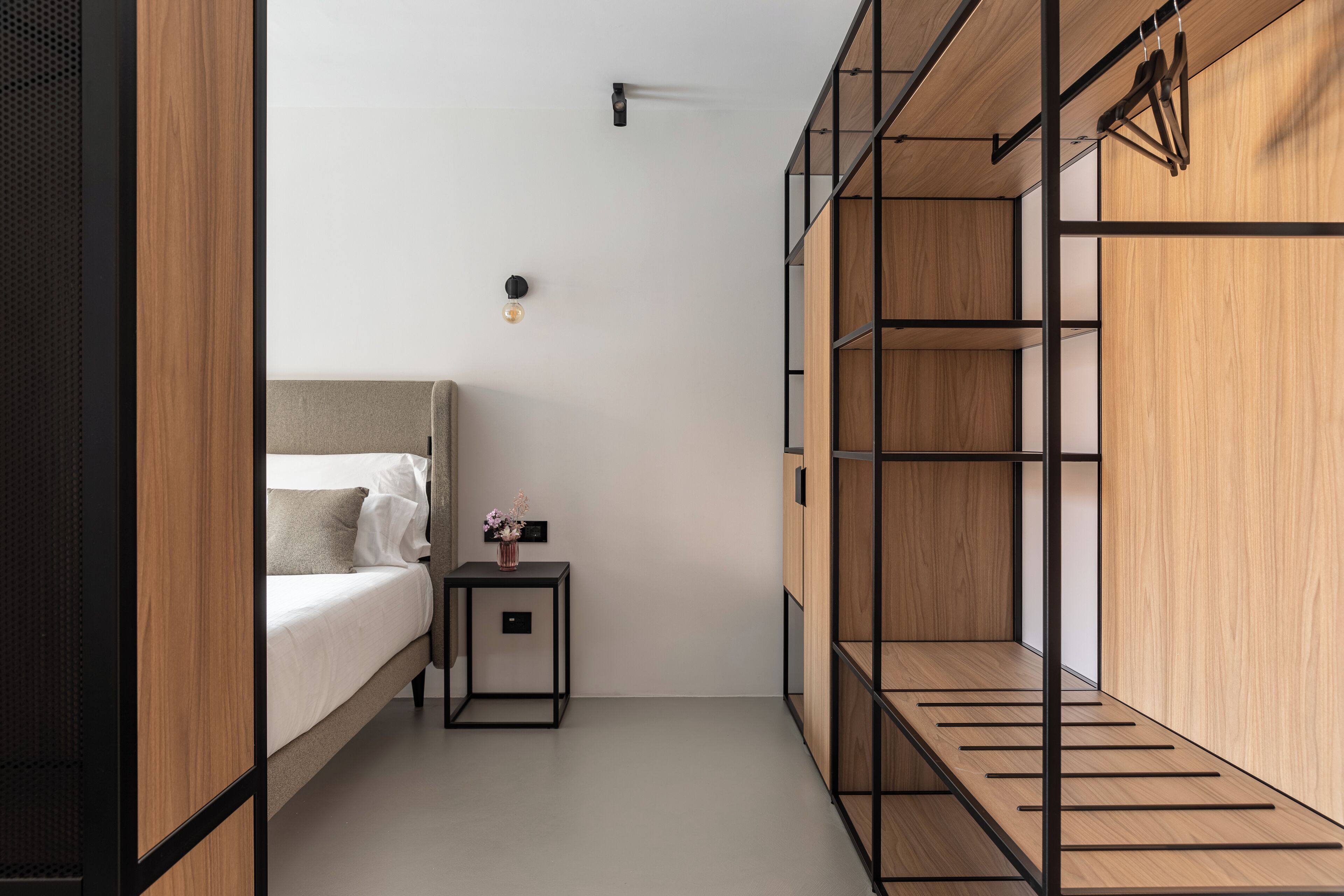 Une chambre à coucher moderne et compacte, comprenant un lit, une petite table de chevet avec un vase, et des étagères en bois intégrées.