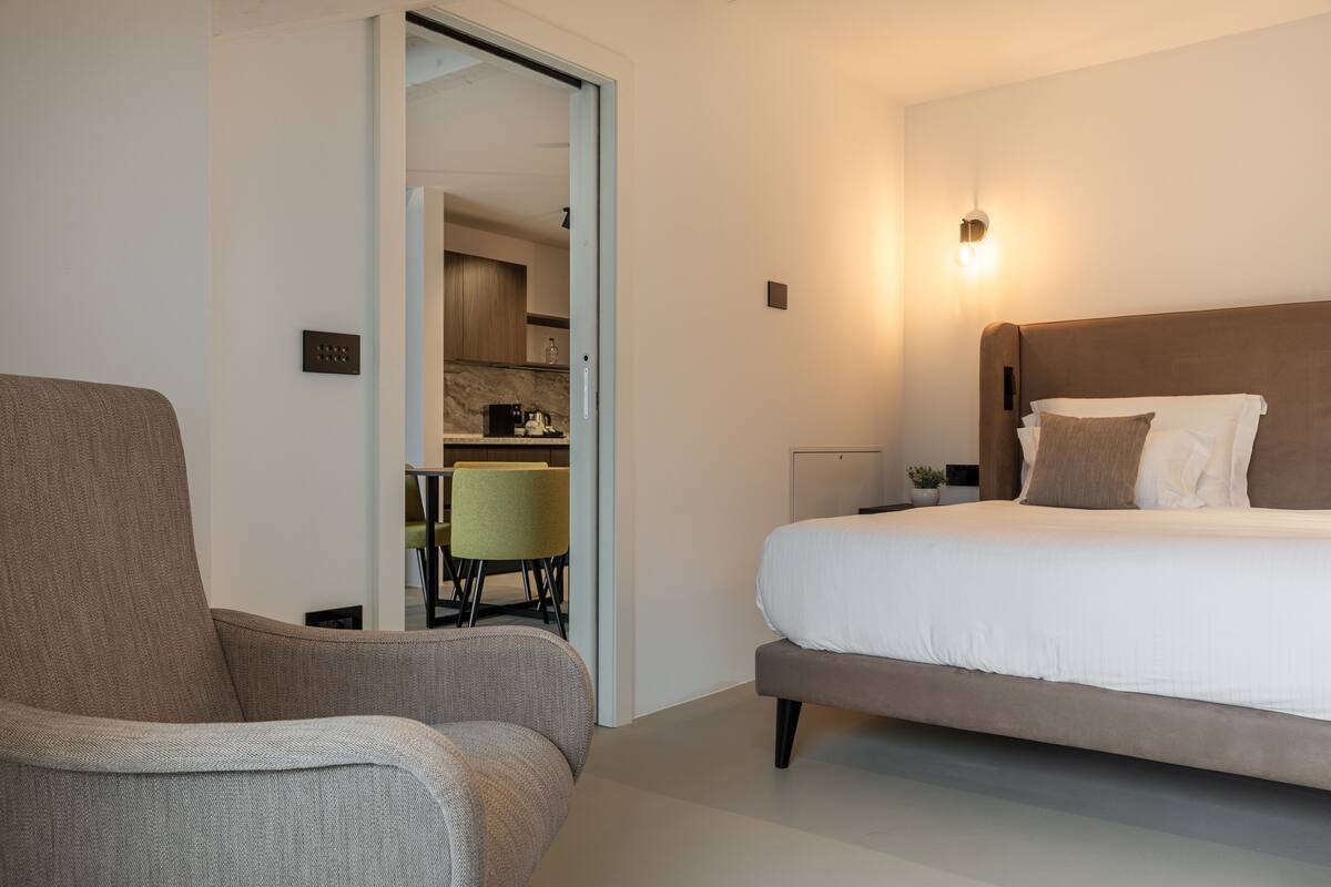 A modern hotel room with a large bed, a sofa, and a kitchenette in the background.