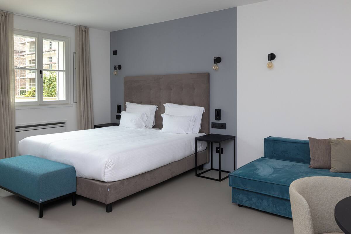 A modern hotel room with a large bed, a small table, a sofa, and a window with curtains.