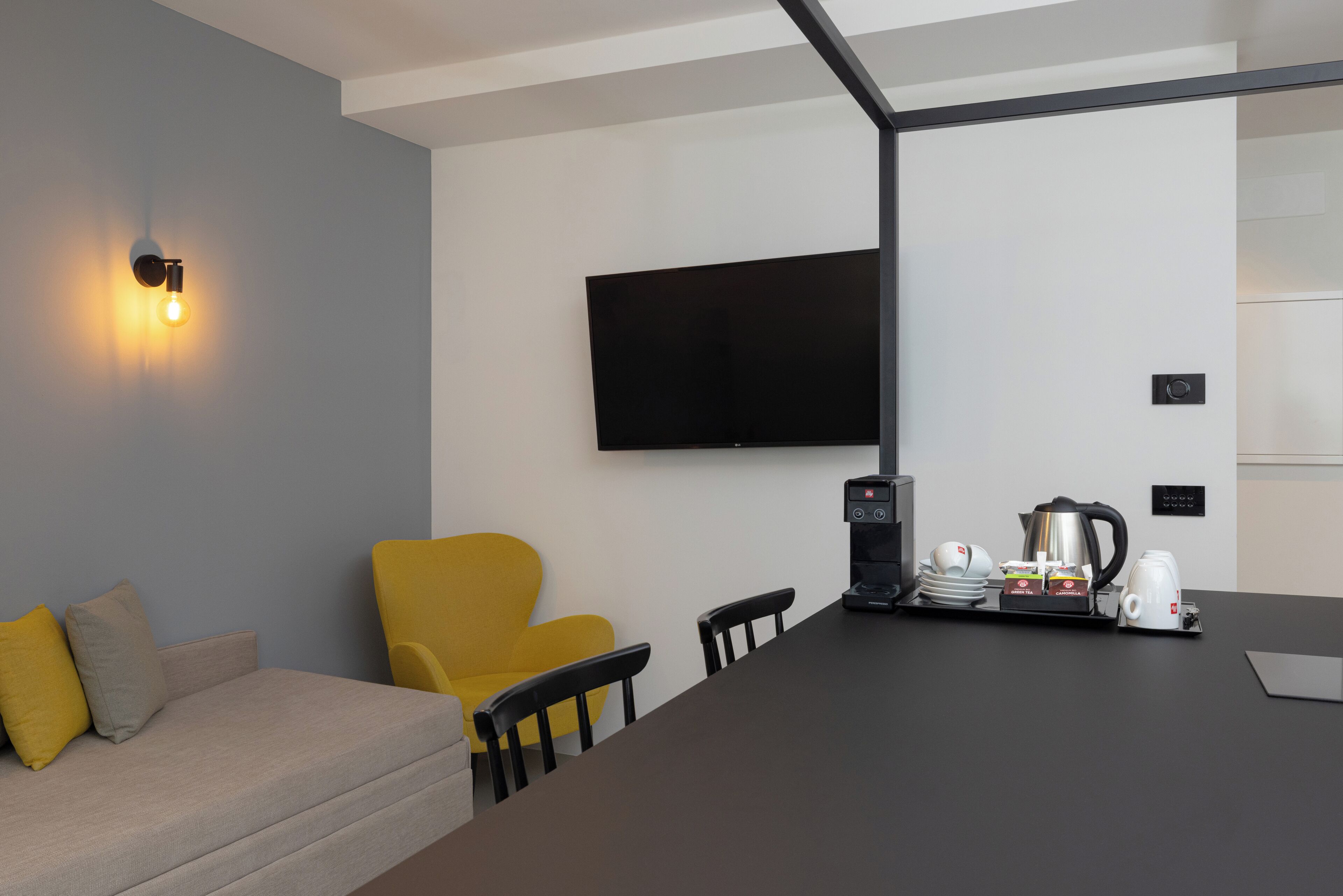 A modern room with a sofa, a yellow armchair, a flat-screen TV mounted on the wall, a coffee station with a coffee maker, cups, and a kettle.