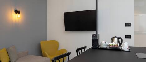 A modern room with a sofa, a yellow armchair, a flat-screen TV mounted on the wall, a coffee station with a coffee maker, cups, and a kettle.