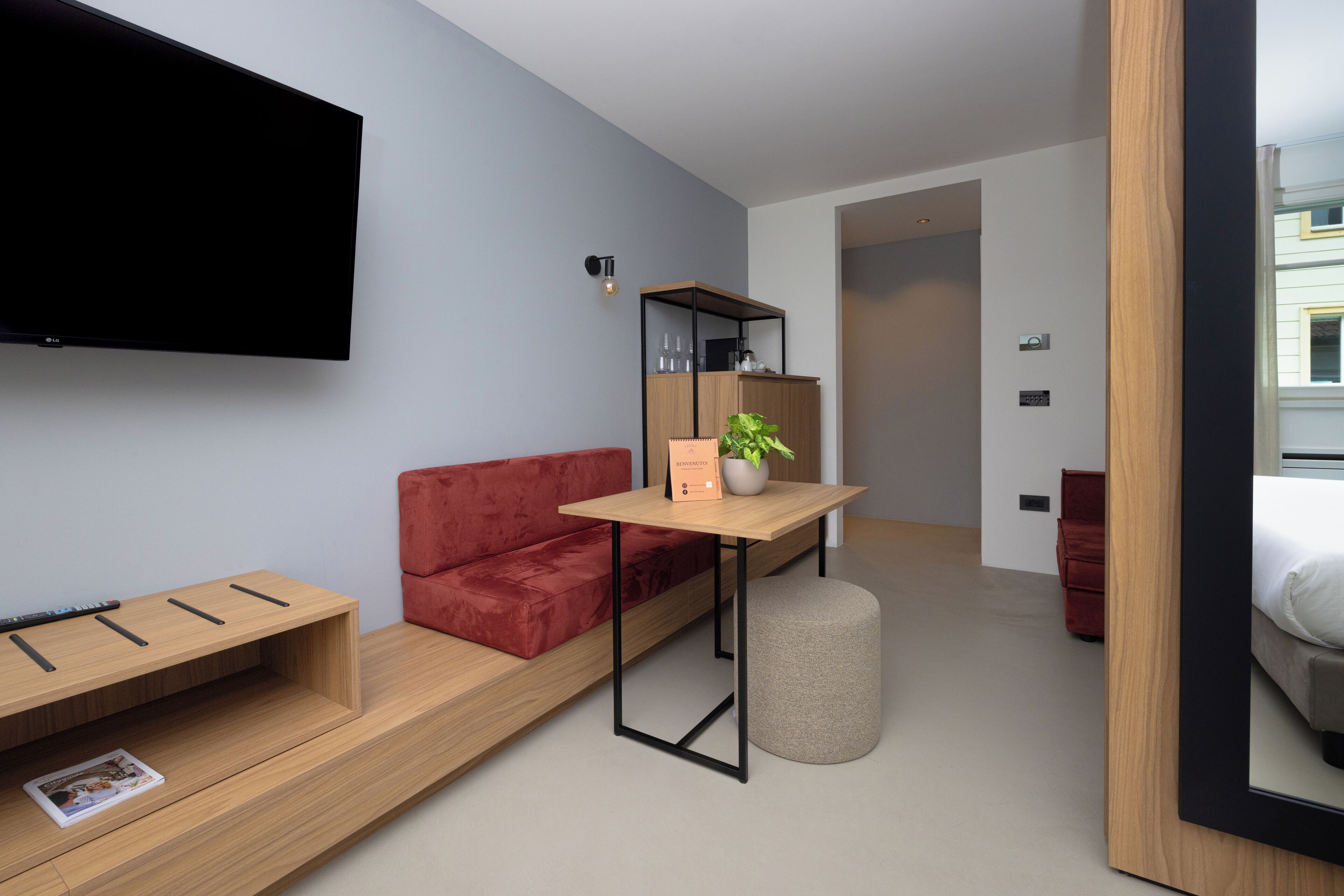A modern hotel room with a flat-screen TV mounted on the wall, a wooden table with a potted plant, a red sofa, and a small ottoman.