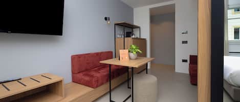 Een moderne hotelkamer met een flatscreen televisie aan de muur, een houten tafel met een potplant, een rode bank en een klein poef.