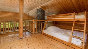 Eine Holzhütte mit Etagenbetten, einem Kamin aus Stein und einem Balkon mit Blick auf Grünflächen.