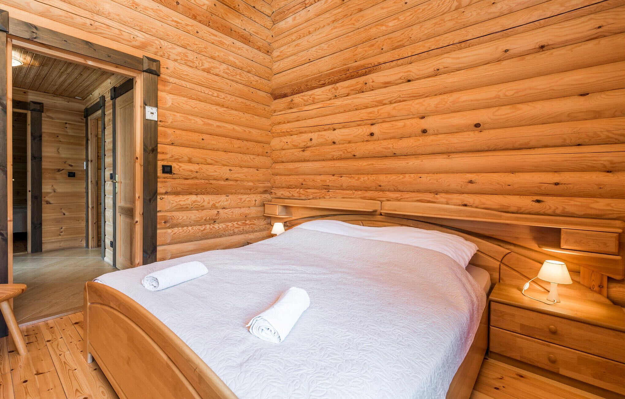 A wooden cabin room with a bed, a nightstand, and a door leading to another room.