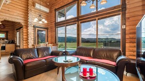 Ein Holzblockhaus-Interieur mit einem Ledersofa, einem Glastisch und großen Fenstern mit Blick auf die Landschaft.