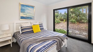 A bedroom with a bed, a nightstand, a lamp, a ceiling fan, a window with a view of greenery, and a painting on the wall. - Aireys Delight (Aireys Inlet)