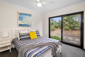 Ein Schlafzimmer mit Bett, Nachttisch, Lampe, Deckenventilator, Fenster mit Blick auf Grünflächen und einem Bild an der Wand.