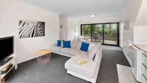 A modern living room with a flat-screen TV, a grey sofa with blue cushions, a small wooden side table, and a large window offering a view of the outdoors. - Aireys Delight (Aireys Inlet)