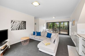 Ein modernes Wohnzimmer mit einem Flachbildfernseher, einem grauen Sofa mit blauen Kissen, einem kleinen Holztisch und einem großen Fenster mit Blick ins Freie.