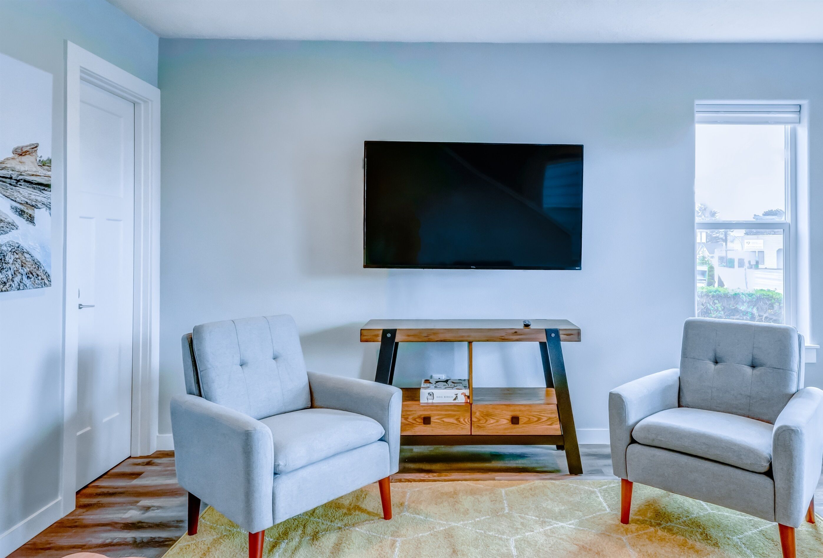 Una sala de estar con dos sillones grises, una mesa de centro de madera y un televisor de pantalla plana montado en la pared.