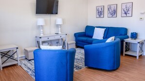 A modern living room with a blue sofa, two armchairs, a TV, and three framed artworks on the wall.