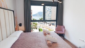 A modern hotel room with a large bed, a view of the city, and a chair by the window. - A016 - ARENAL SUITES - AVANOA (Calpe)
