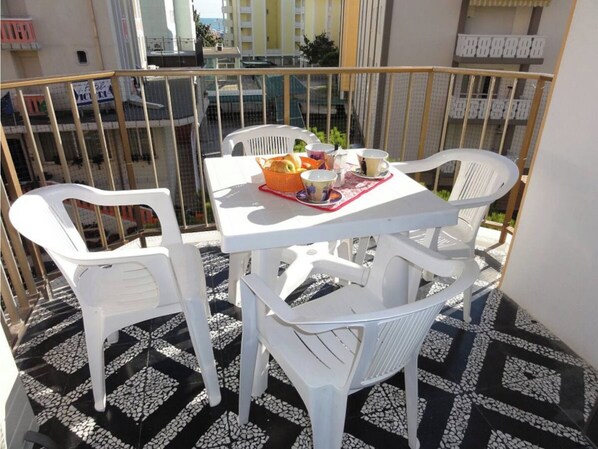 A balcony with a white table and chairs, a tray with cups and a basket of bread, and a tiled floor.