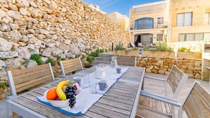 Una mesa de madera dispuesta con frutas, tazas y una jarra, al aire libre con muros de piedra y un edificio en el fondo.