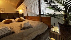 Ein Schlafzimmer mit Holzboden, einem Bett mit weißer und brauner Bettwäsche, einer großen Kübelpflanze und einem kleinen Tisch mit einer Lampe.