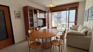 A dining area with a wooden table and chairs, a built-in bookshelf, and a balcony with a view. - Lido dei Pini three-room apartment sleeps 6 (Bibione)