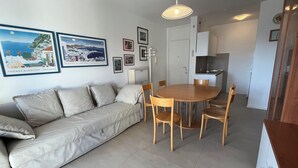 A modern living room with a beige sofa, wooden dining table, and chairs. There are framed pictures on the wall and a kitchen area with cabinets and a sink. - Lido dei Pini three-room apartment sleeps 6 (Bibione)