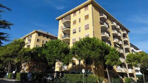 A multi-story residential building with balconies, surrounded by trees and a clear blue sky. - Lido dei Pini three-room apartment sleeps 6 (Bibione)
