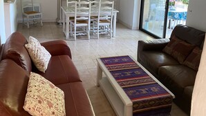 Un salon avec un canapé en cuir marron, une table basse à motifs et un fauteuil en cuir marron. On y trouve également un coin repas avec des meubles blancs et des luminaires suspendus.