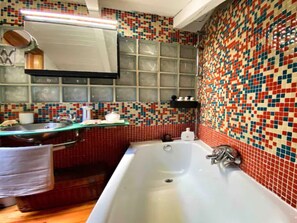 Ein Badezimmer mit einer großen Badewanne, einem Waschbecken und einem Spiegel. Die Wände sind mit einem farbenfrohen Mosaikmuster gefliest.