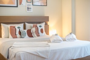 A neatly made bed with white linens and decorative pillows, a bedside lamp, and a folded towel on the nightstand. - Supreme Suite with balcony by Cloudkeys (Athens)