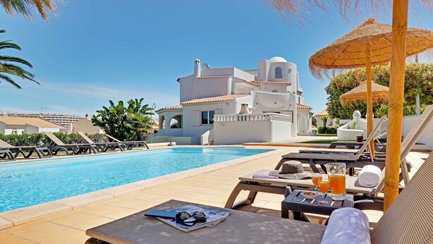 A poolside area with lounge chairs, a table with a towel, and a building in the background. - Villux - Villa Enar - 6 rooms, ideal for families. (Albufeira)