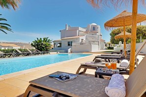 A poolside area with lounge chairs, a table with a towel, and a building in the background. - Villux - Villa Enar - 6 rooms, ideal for families. (Albufeira)