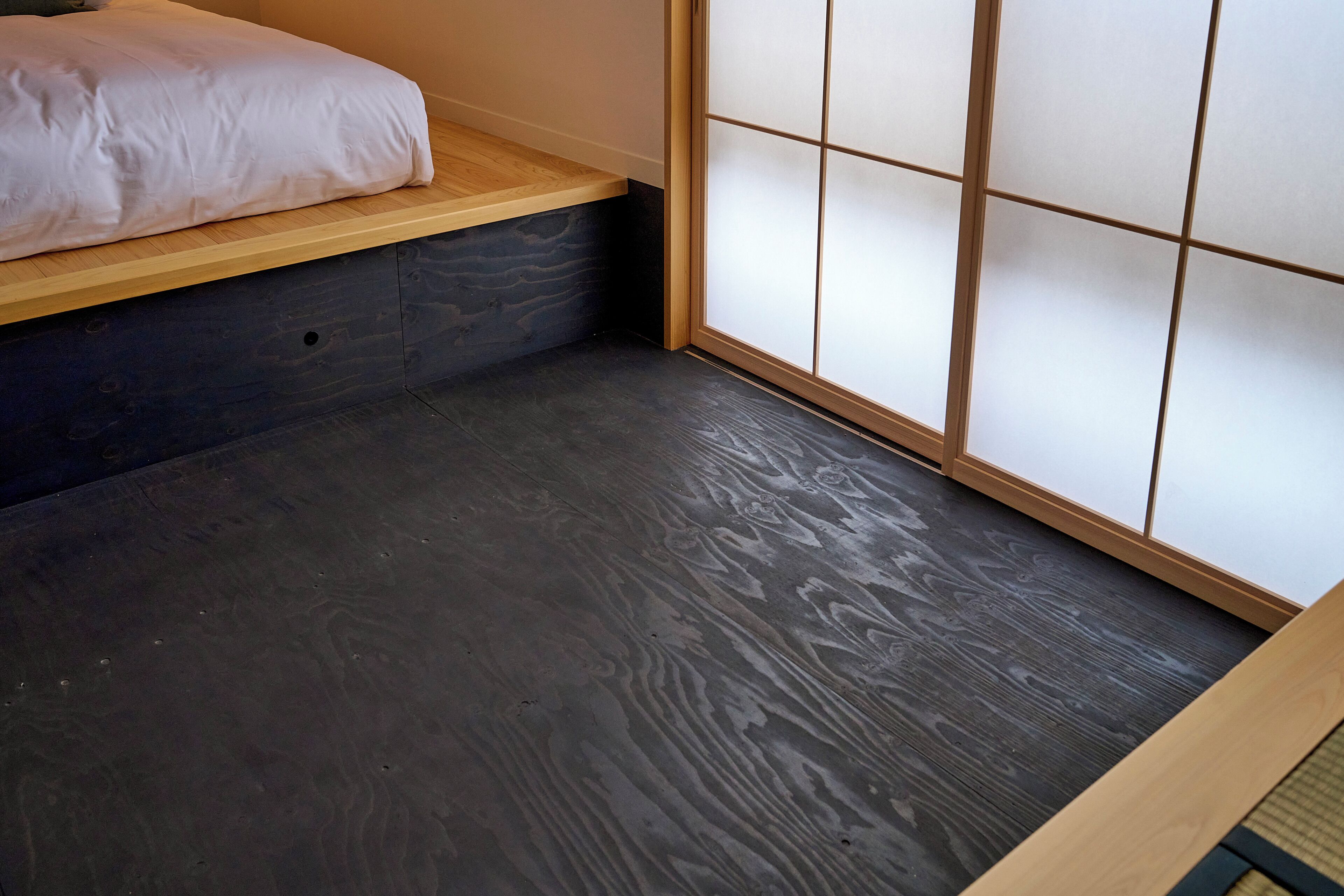 Una habitación con una cama sobre una plataforma de madera, piso de madera oscura y una puerta corredera tradicional con paneles de vidrio esmerilado.