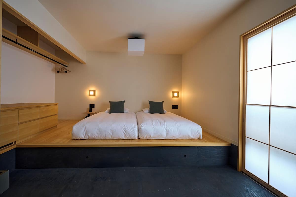 Une chambre moderne avec un lit surélevé en bois, deux tables de chevet, des lampes fixées au mur et une porte coulissante de type shoji.
