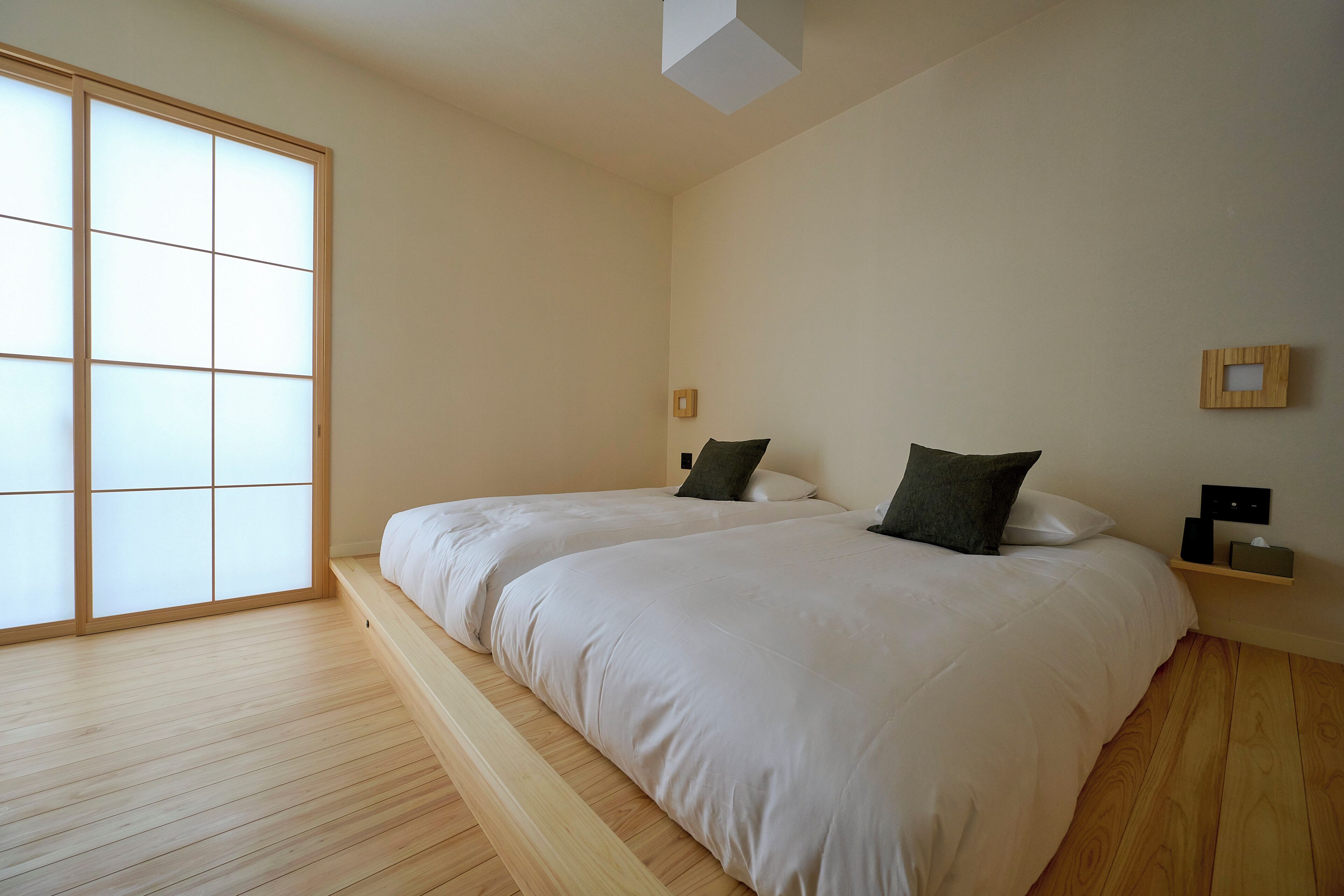 Ein schlicht eingerichtetes Schlafzimmer mit zwei Betten, einem verschiebbaren Shoji-Bildschirm, Holzboden und einem kleinen Regal mit Lampe und Taschentuchbox.