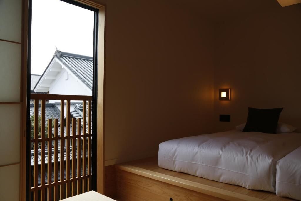 A bedroom with a bed, a window with a view of buildings, and a small wall-mounted light.