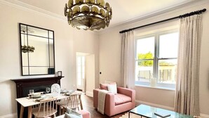 A living room with a pink sofa, a glass coffee table, a fireplace, a dining area, and a large window with curtains.