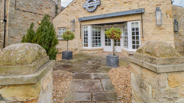 A stone building with a sign reading "The Grage Bridge." It features a stone entrance, potted plants, and a stone pathway. - Burns Cottage (Barnard Castle)