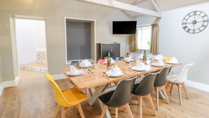 A dining room with a long table set for a meal, surrounded by chairs, a TV mounted on the wall, and a clock. - Burns Cottage (Barnard Castle)