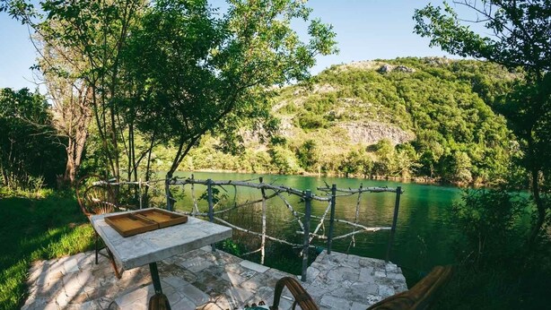 Eine Steinterrasse mit Tisch und Stühlen, die auf einen stillen See mit üppiger Vegetation und Hügeln blickt.