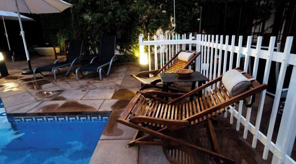 Zona de piscina con sillas de descanso y una mesa, rodeada por una valla blanca.