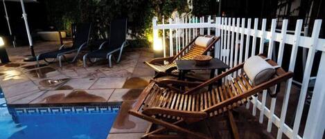 Zona de piscina con sillas de descanso y una mesa, rodeada por una valla blanca.