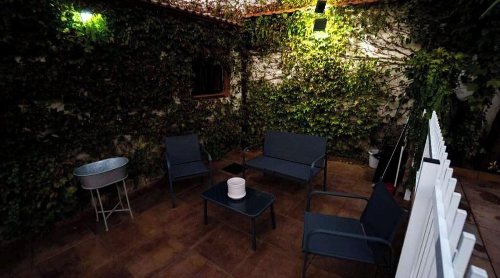 Un patio al aire libre con suelo de piedra rústica, rodeado de abundante vegetación y muros cubiertos de hiedra. Hay dos sillas de color oscuro y una mesa pequeña con un objeto blanco encima.