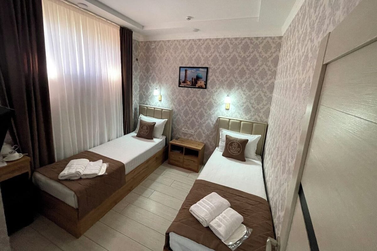 Habitación de hotel con dos camas individuales, una mesita de noche de madera, un cuadro colgado en la pared y una ventana con cortinas.