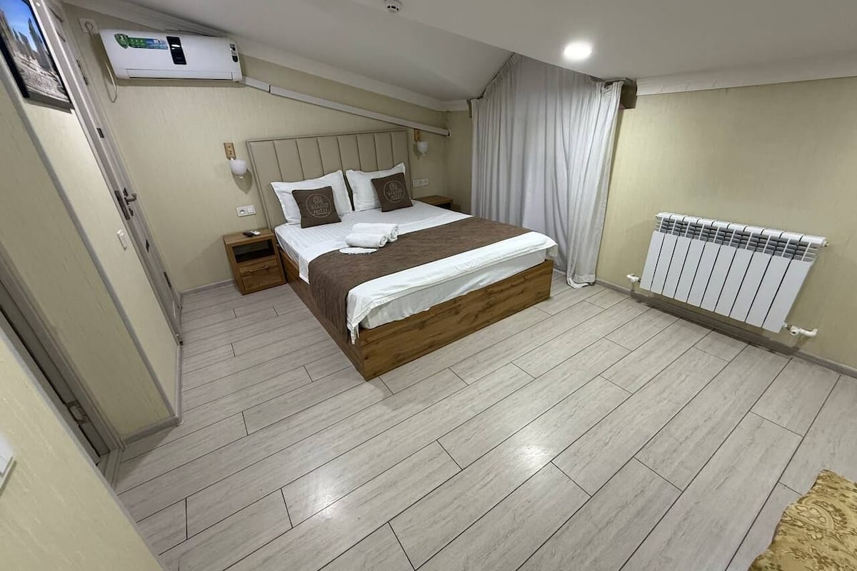 A bedroom with a large bed, a wooden headboard, a bedside table, a nightstand, an air conditioning unit, a radiator, and a curtain.