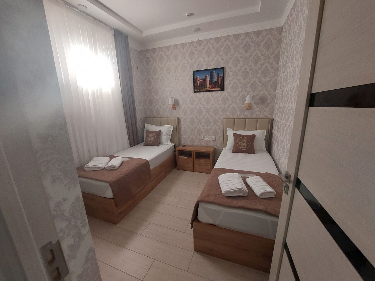 A hotel room with two beds, a small wooden nightstand, a wall-mounted TV, and a patterned wallpaper.
