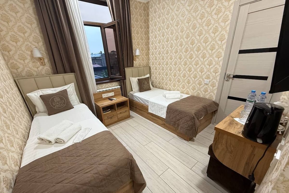 A hotel room with two beds, a wooden nightstand, a desk with a coffee maker, and a view of the city through the window.