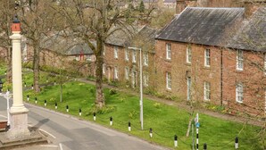 A street view with a row of brick houses, a white monument, and a grassy area with yellow flowers. - South Lodge (Appleby-in-Westmorland)