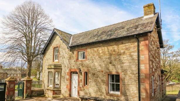 A stone building with a traditional design, featuring a central door and windows, surrounded by a paved area and a tree. - South Lodge (Appleby-in-Westmorland)